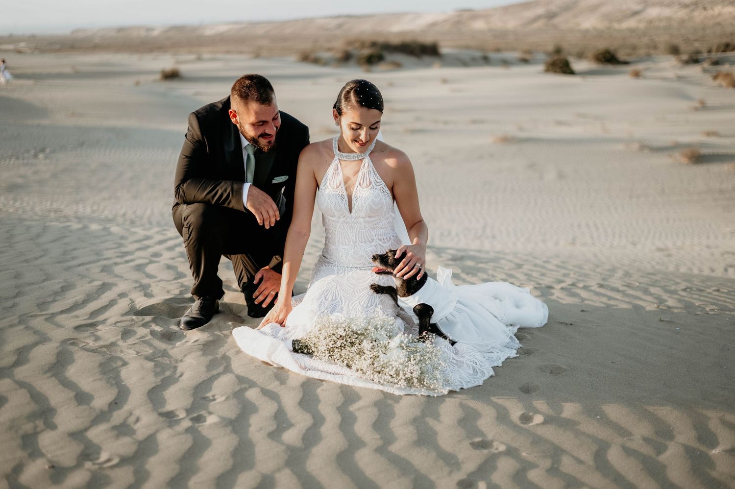 A bride and groom kneel together on rippling sand dunes in a desert landscape during their wedding photoshoot.