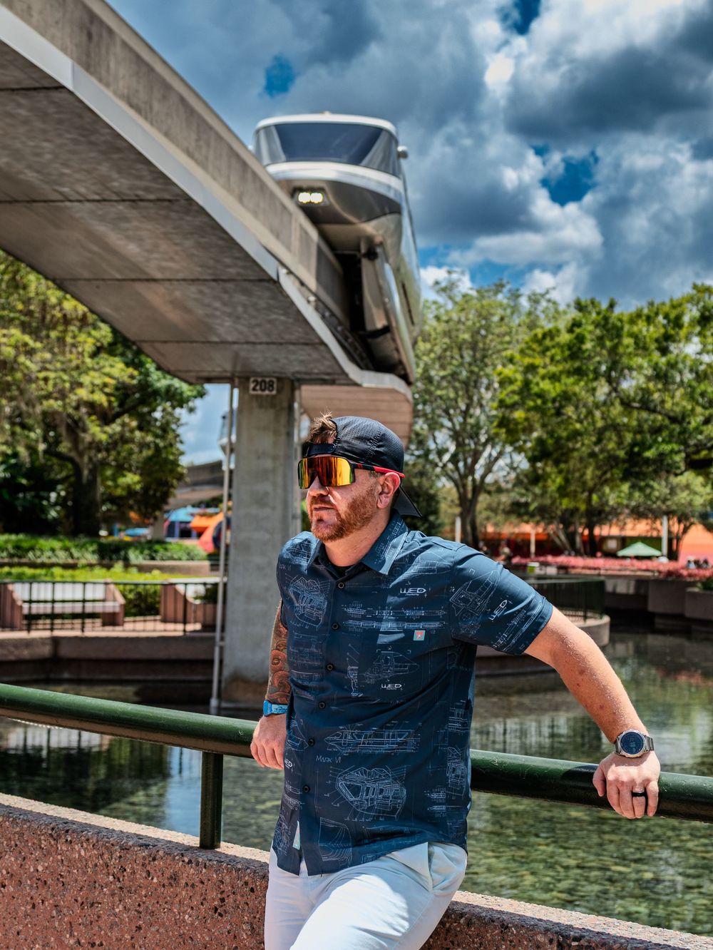 Person in denim shirt and sunglasses poses near water with dramatic cloudy sky in background.