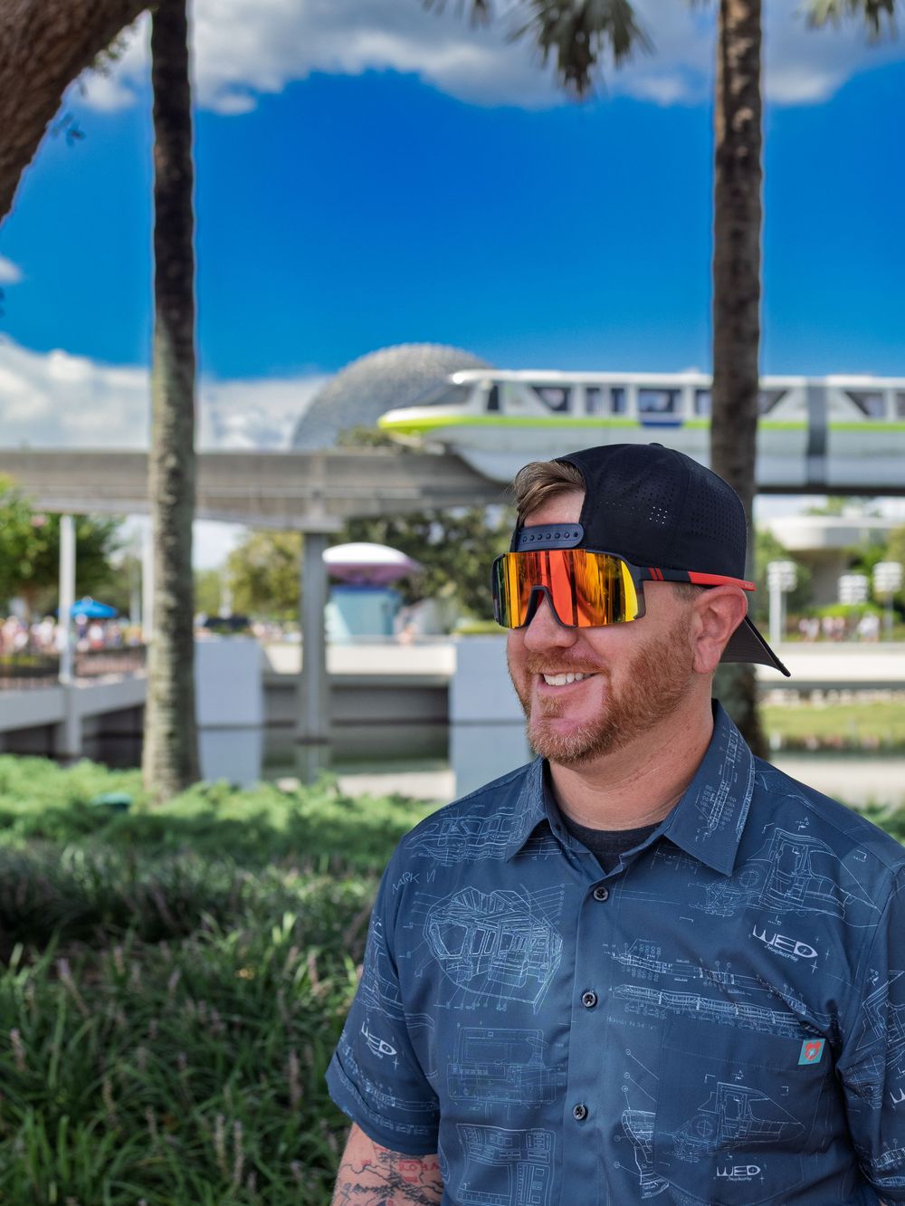 Person wearing sunglasses and denim shirt poses outdoors with palm trees and blue sky.