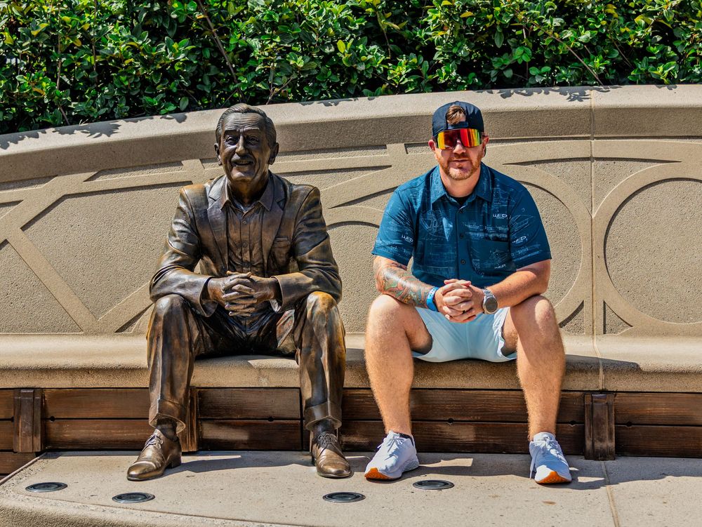 Person sitting next to bronze statue on bench with lush greenery in background.