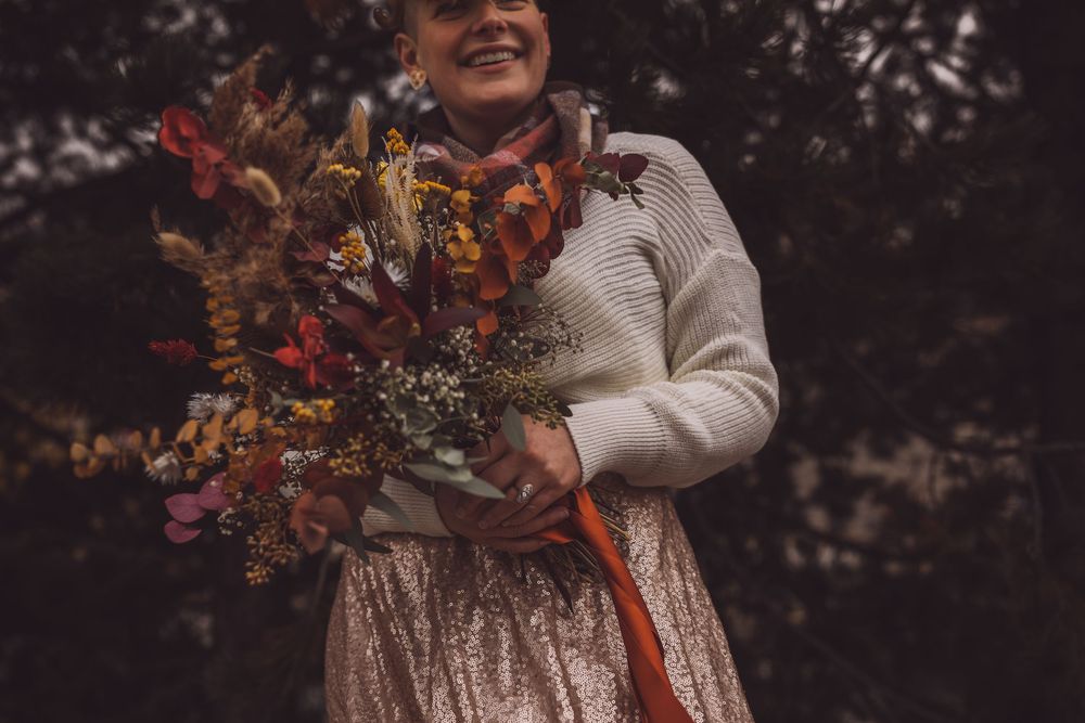 A person in beige clothing sits outdoors holding vibrant autumn-colored flower arrangements against a dark background.