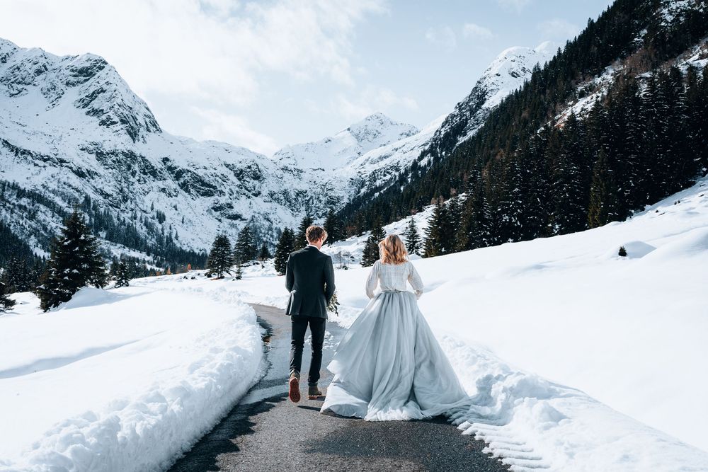 A couple in winter wedding attire walks down a snowy mountain path surrounded by snow-capped Alpine peaks.