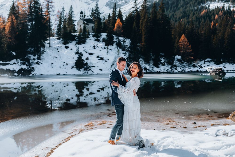A couple in winter wedding attire embraces on a snowy lakeshore with evergreen trees and mountains in the background.
