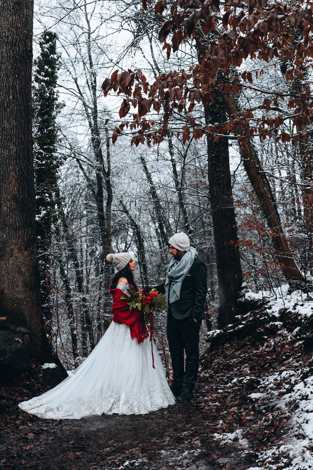 A couple in winter wedding attire stands on a snowy forest path surrounded by bare trees and fallen autumn leaves.