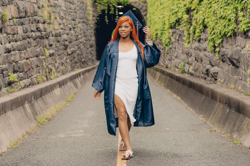 High school graduate in blue gown and white dress walking down concrete path between stone walls in Alexandria, VA