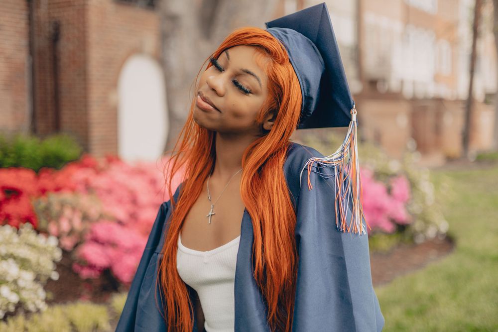 Graduate in blue cap and gown with vibrant orange hair stands near blooming azalea bushes during commencement day.