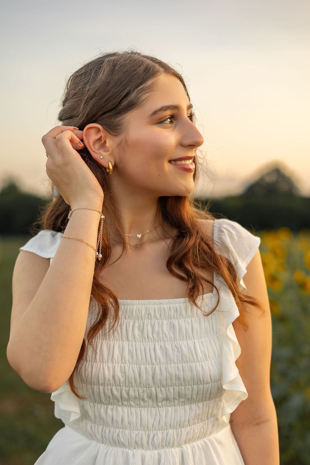 Young woman in white ruffled summer dress smiles while adjusting her hair during golden hour in a flower field.