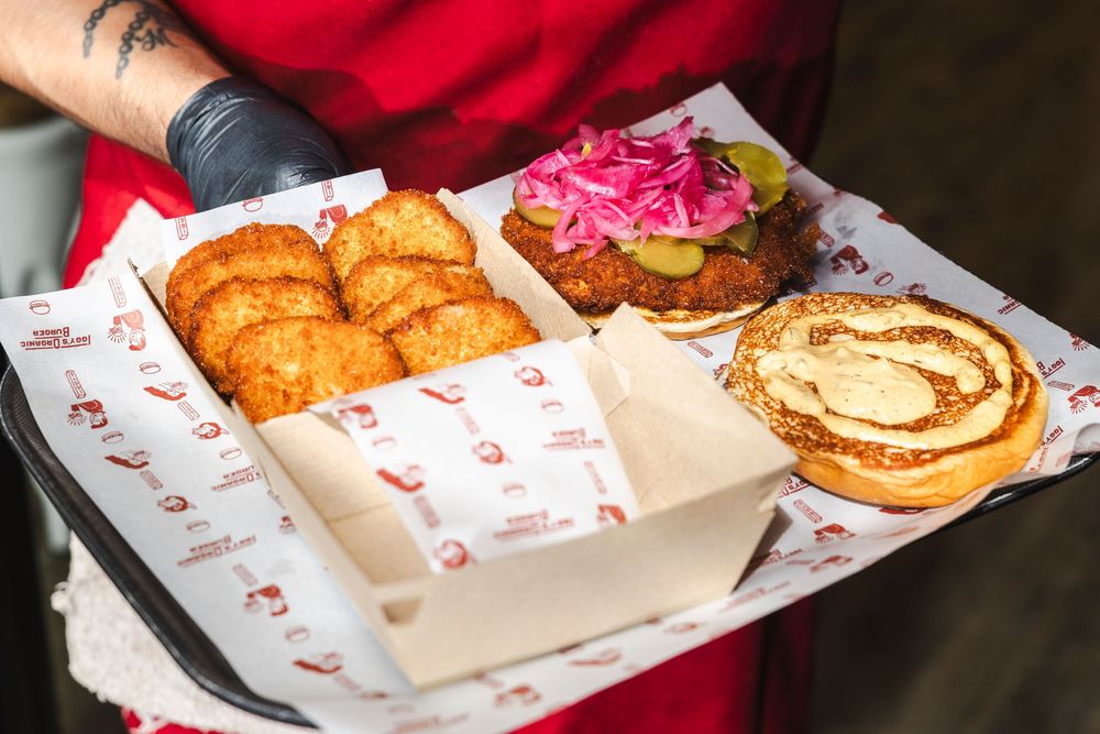 Two hands holding a brown paper bag filled with assorted food items from a restaurant on a red surface.