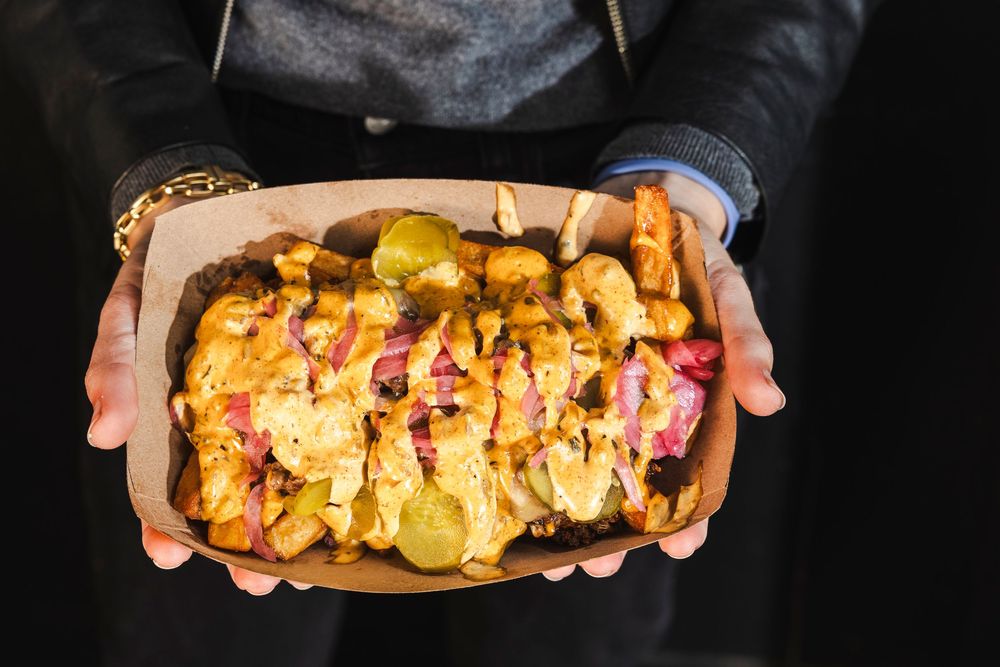 Hands holding a cardboard box filled with loaded potato chips topped with cheese, bacon, and red onions.