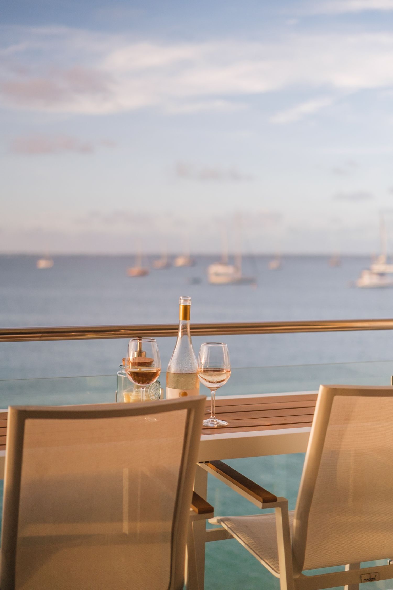 Wine glasses and decanter on balcony railing overlooking peaceful harbor with boats at sunset.