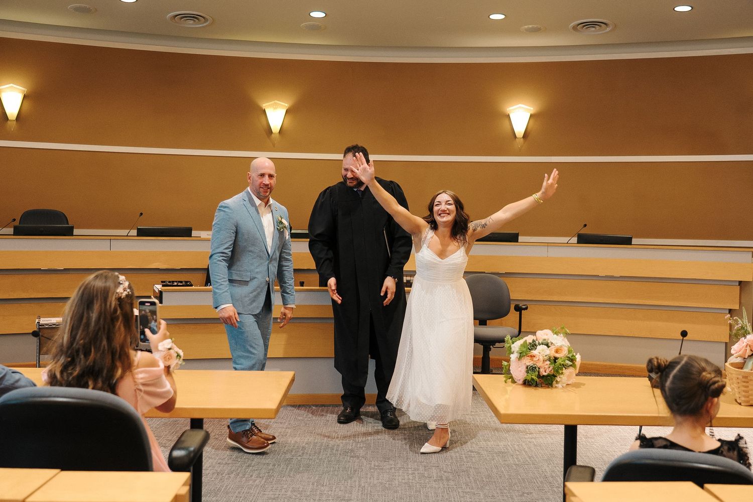 A celebratory moment in a lecture hall with three people posing together and floral arrangements on display.