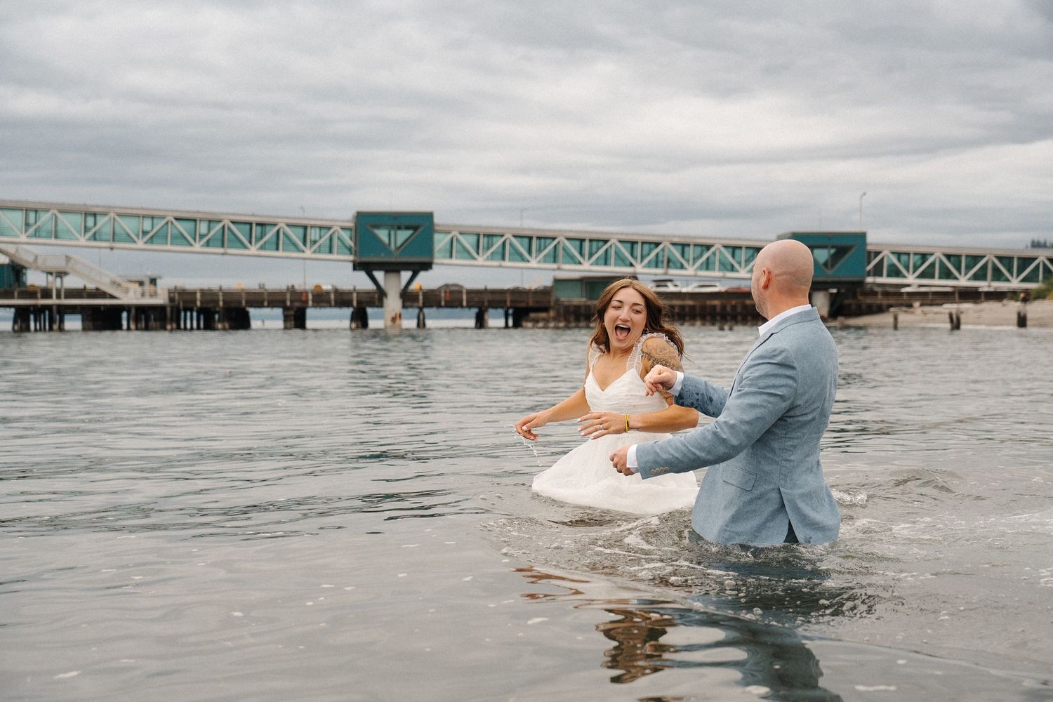 A couple in wedding attire playfully splash in the ocean water near a green bridge structure on an overcast day.