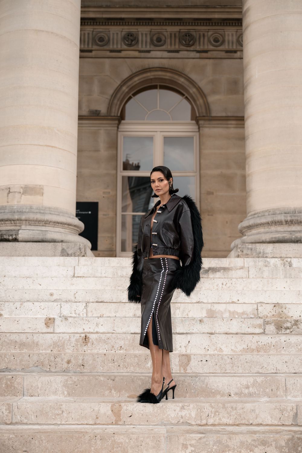 Fashionable outfit featuring black fur coat and pencil skirt on grand stone steps of classical building entrance.