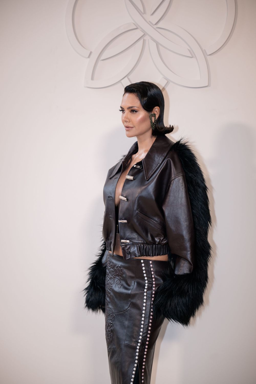 Model wearing edgy black leather outfit with furry sleeves against white wall with decorative design.