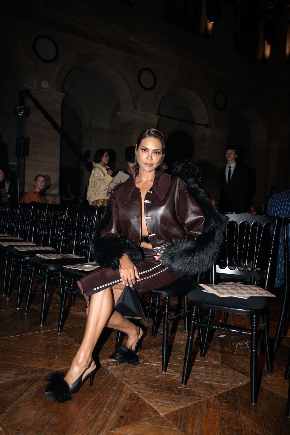 A fashion show attendee wearing a brown leather outfit and black fur accessory sits elegantly in a dark venue.