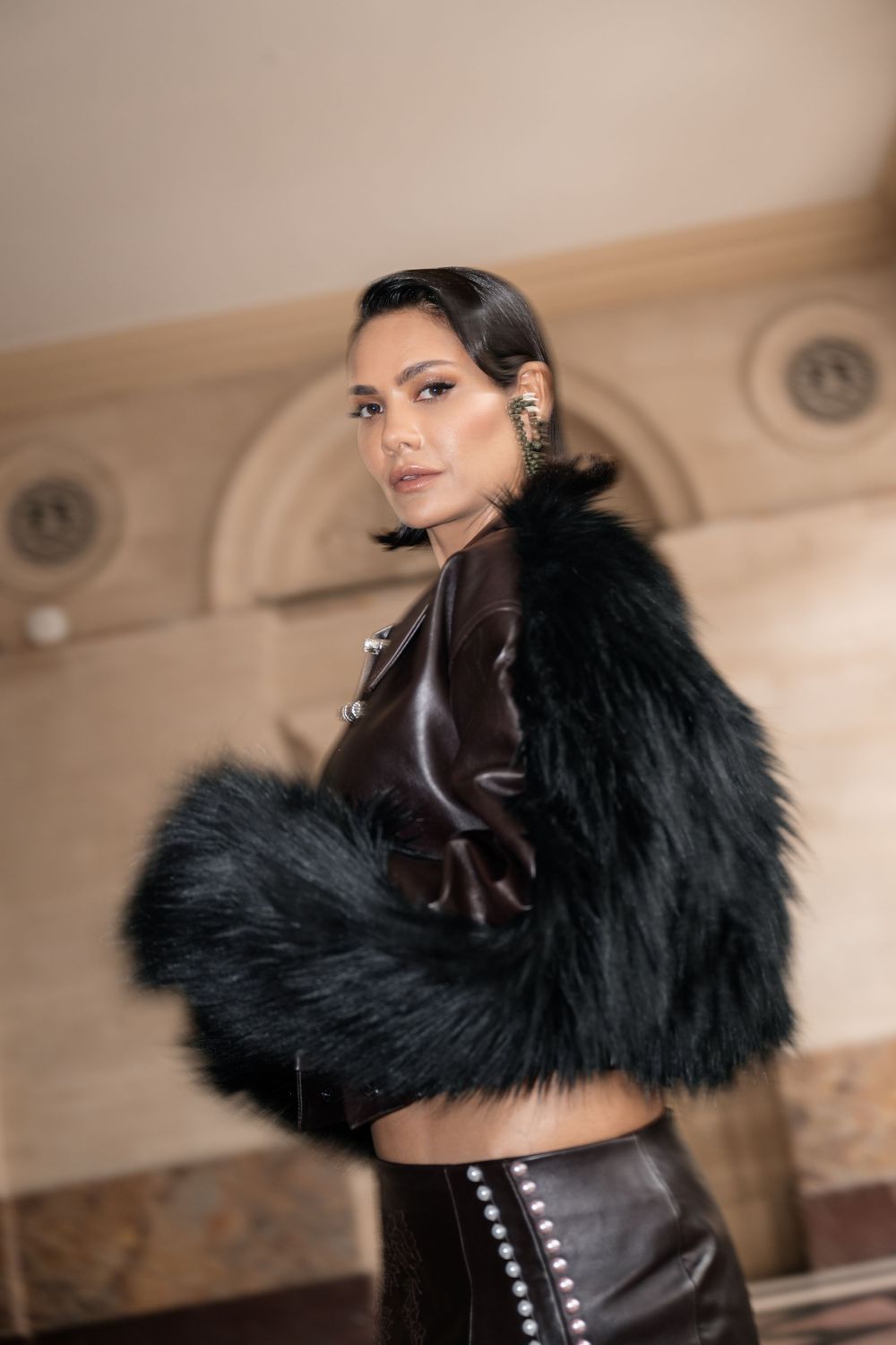 Fashion model wearing black fur coat and leather outfit poses elegantly against neutral interior background.