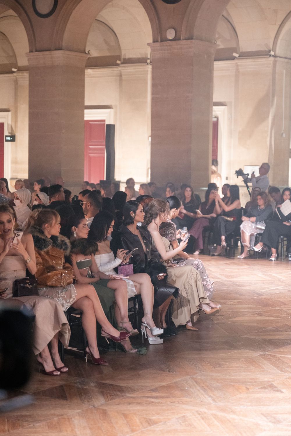 Fashion show audience seated in elegant arched venue with parquet flooring.