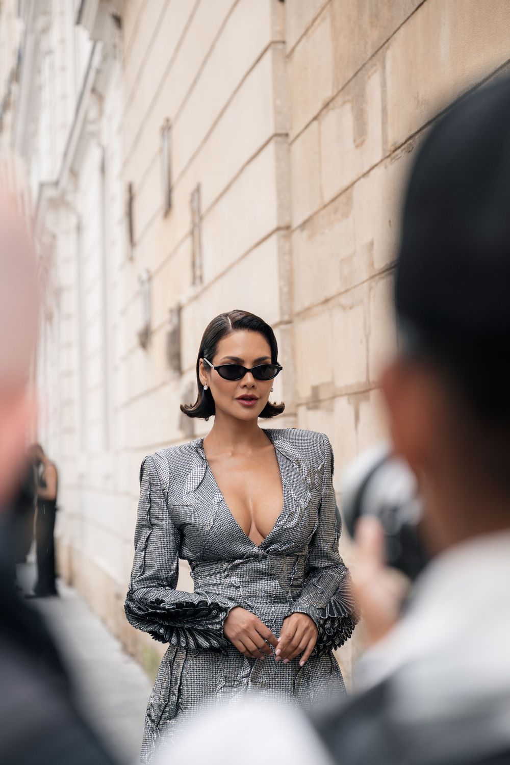 Elegant model in structured grey blazer dress and sunglasses poses against stone wall outdoors.