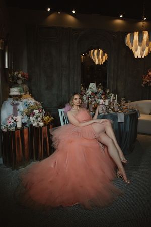 A glamorous scene with someone in a flowing peach tulle gown seated in an elegant room with floral arrangements and chandeliers.