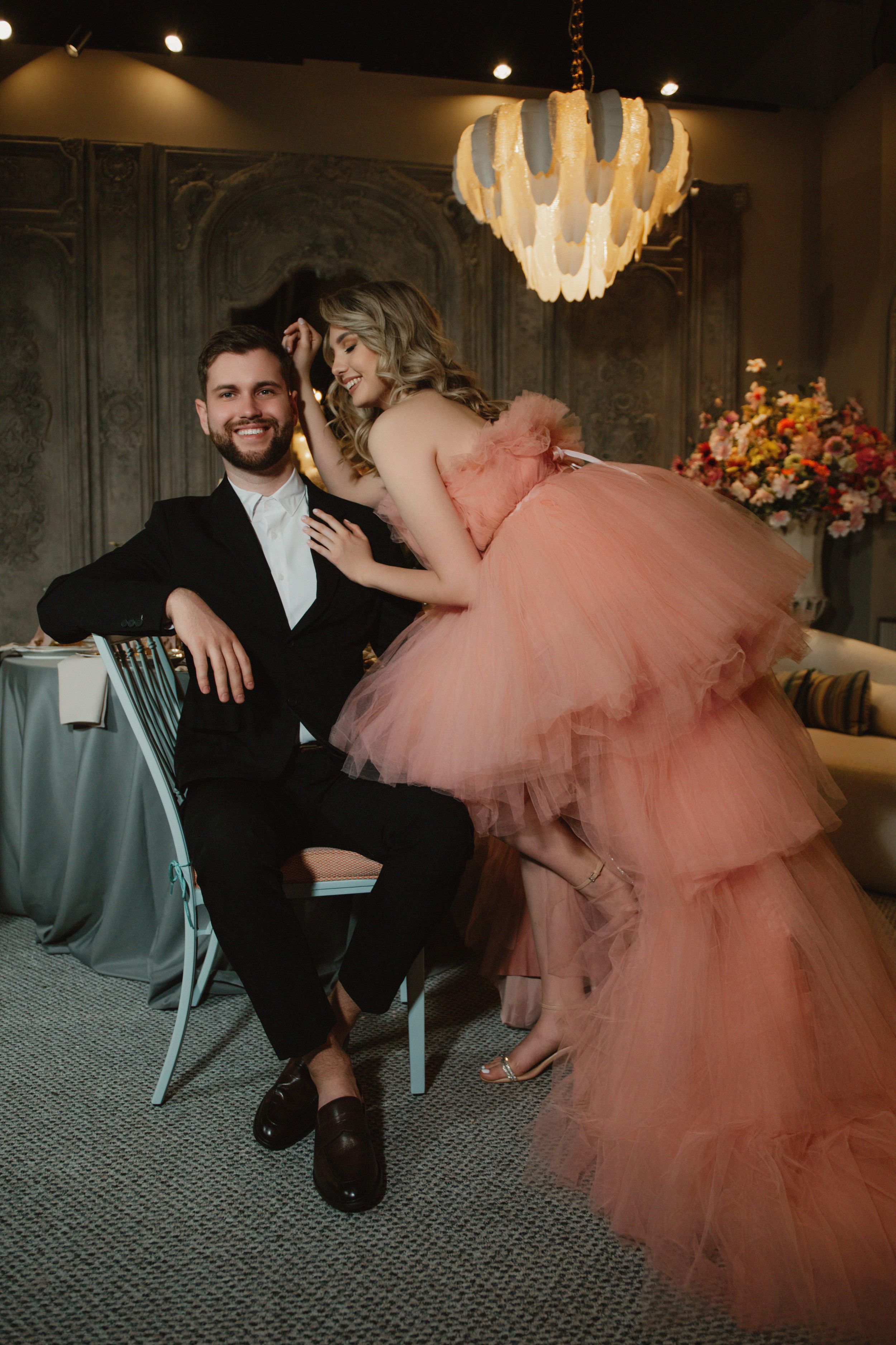 Elegant couple poses in formal attire with peach tulle gown and tuxedo under decorative chandelier at event venue.