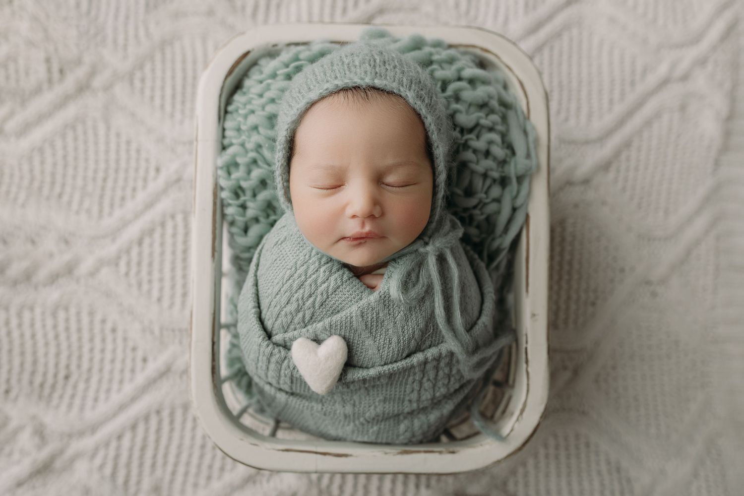 A sleeping baby wrapped in soft sage green knit fabric lies peacefully in a white basket with a heart accent.