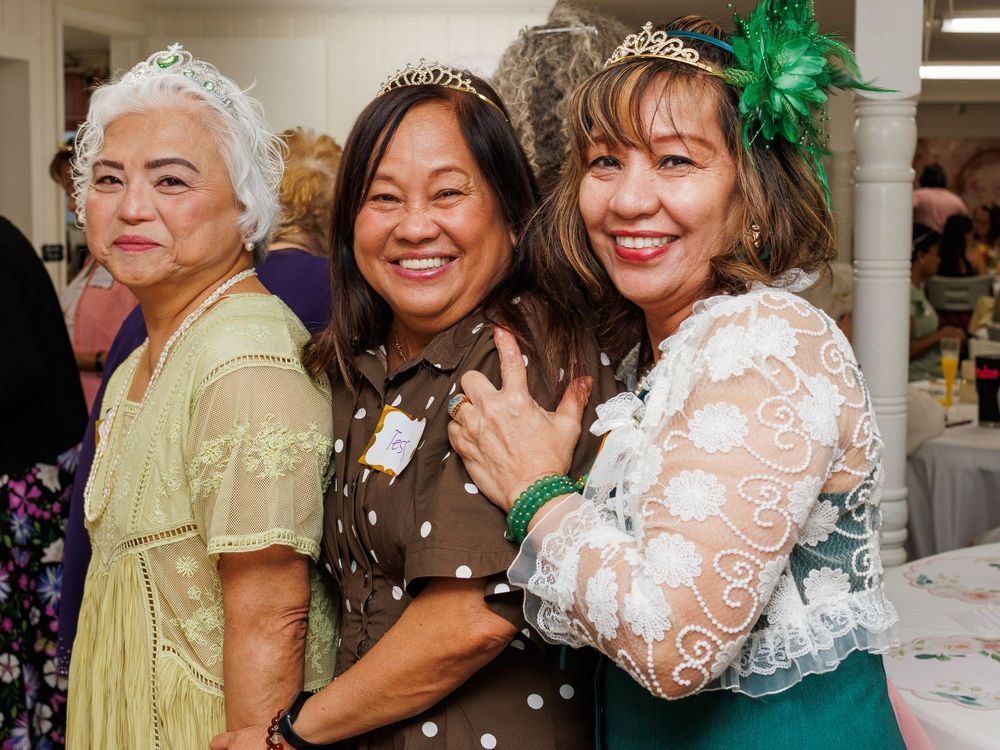 Three women wearing tiaras smile and pose together at a festive indoor event, dressed in elegant green and brown outfits.