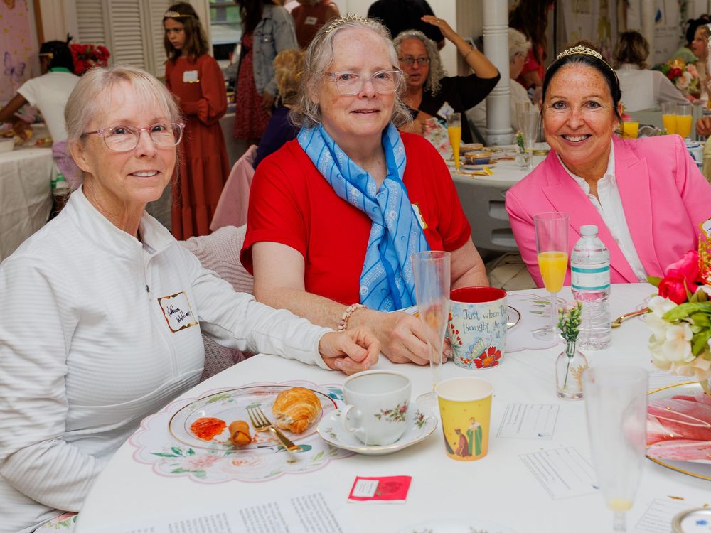 Three women smile together at a decorated banquet table during a formal luncheon event.