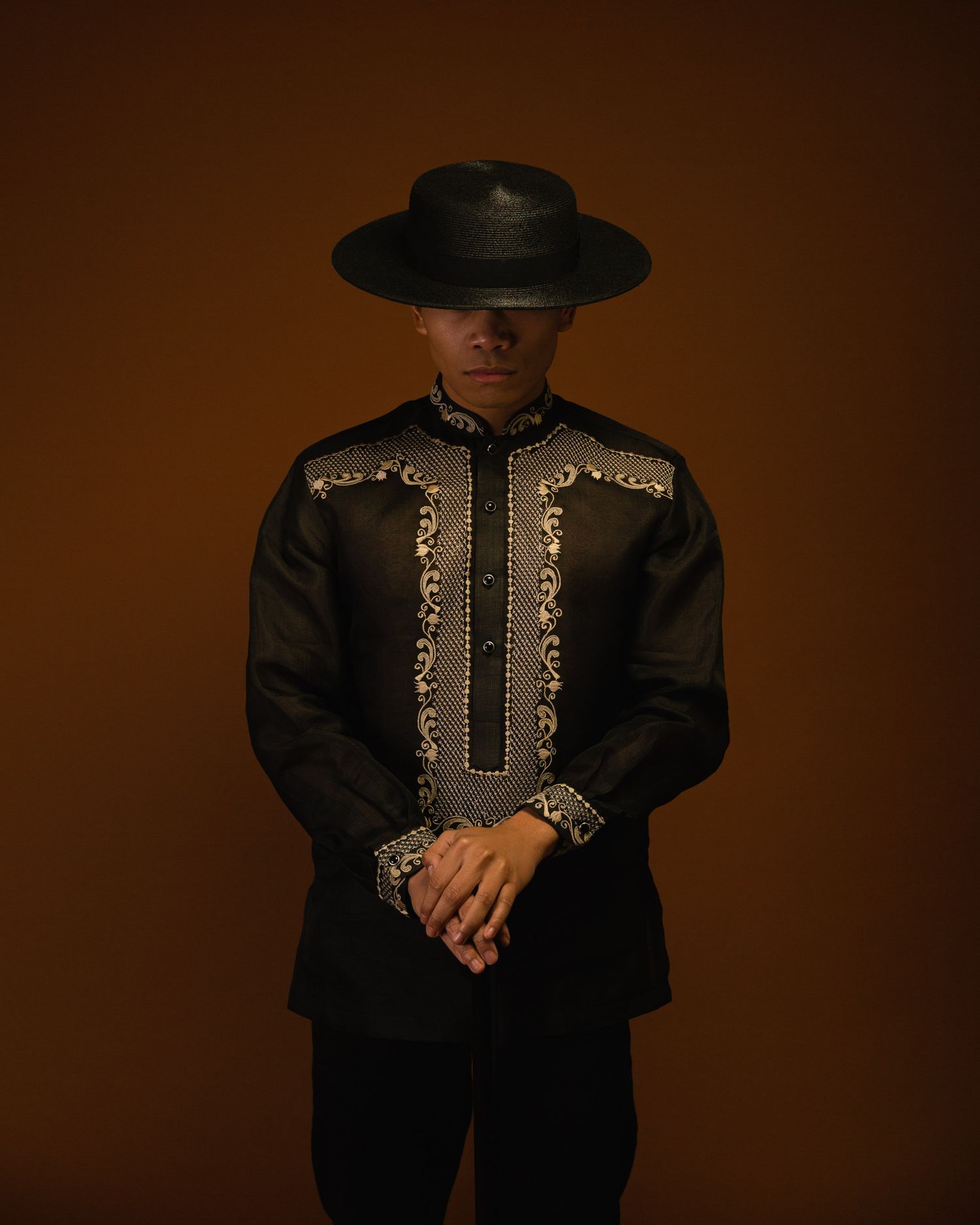 Figure in black Western-style embroidered jacket and hat stands against brown background in dramatic studio lighting.