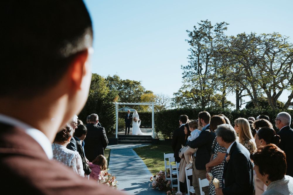 Guests watch wedding ceremony outdoors in formal attire during golden hour at scenic outdoor venue.