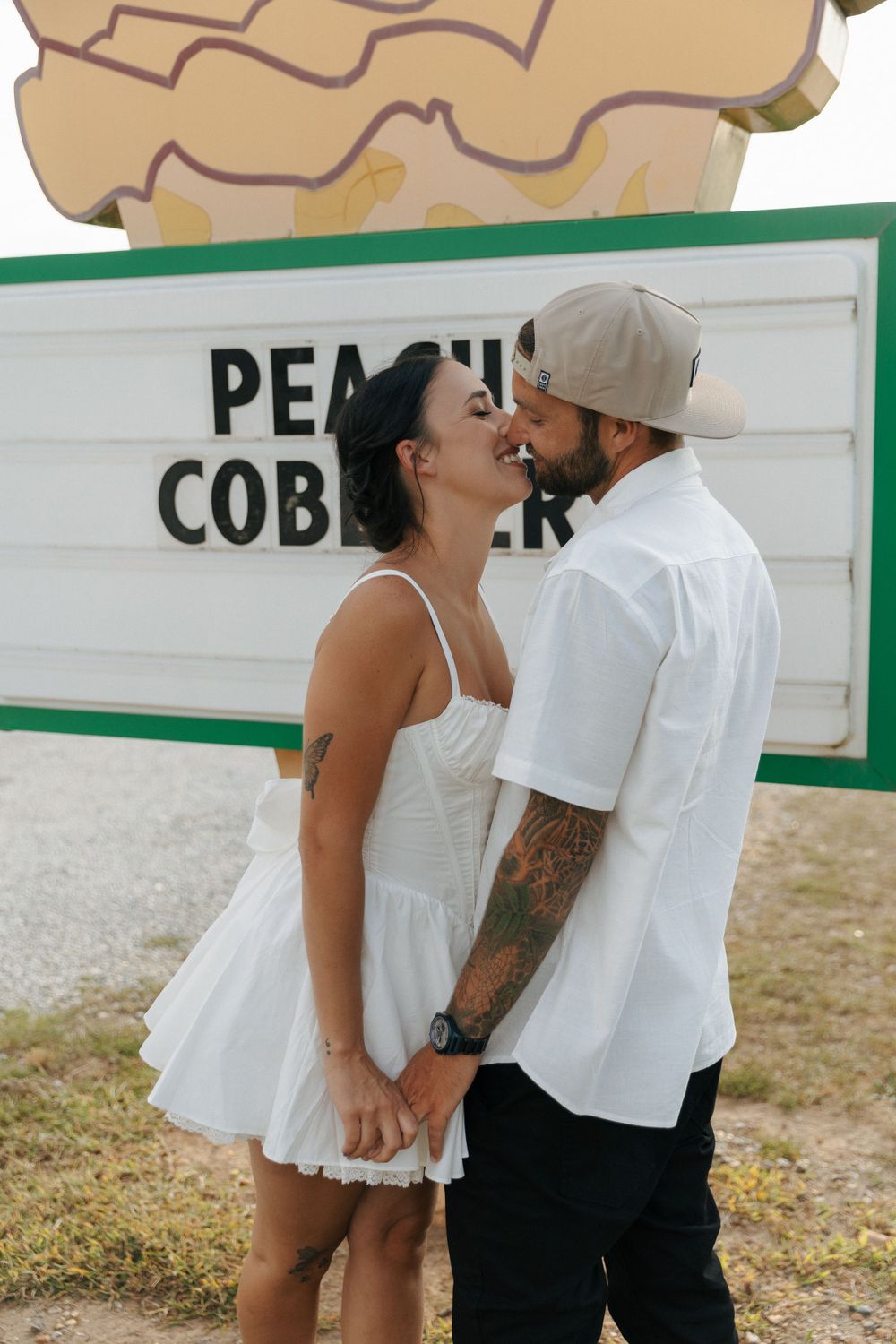 A couple sharing a tender kiss in front of a rustic sign, with the person in white wearing a flowy summer dress.