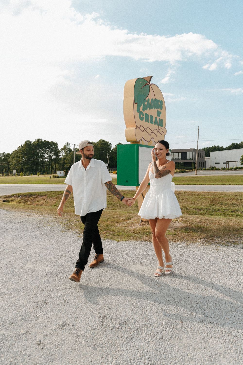 A couple walking together on a gravel path near a large vintage-style sign on a sunny day.