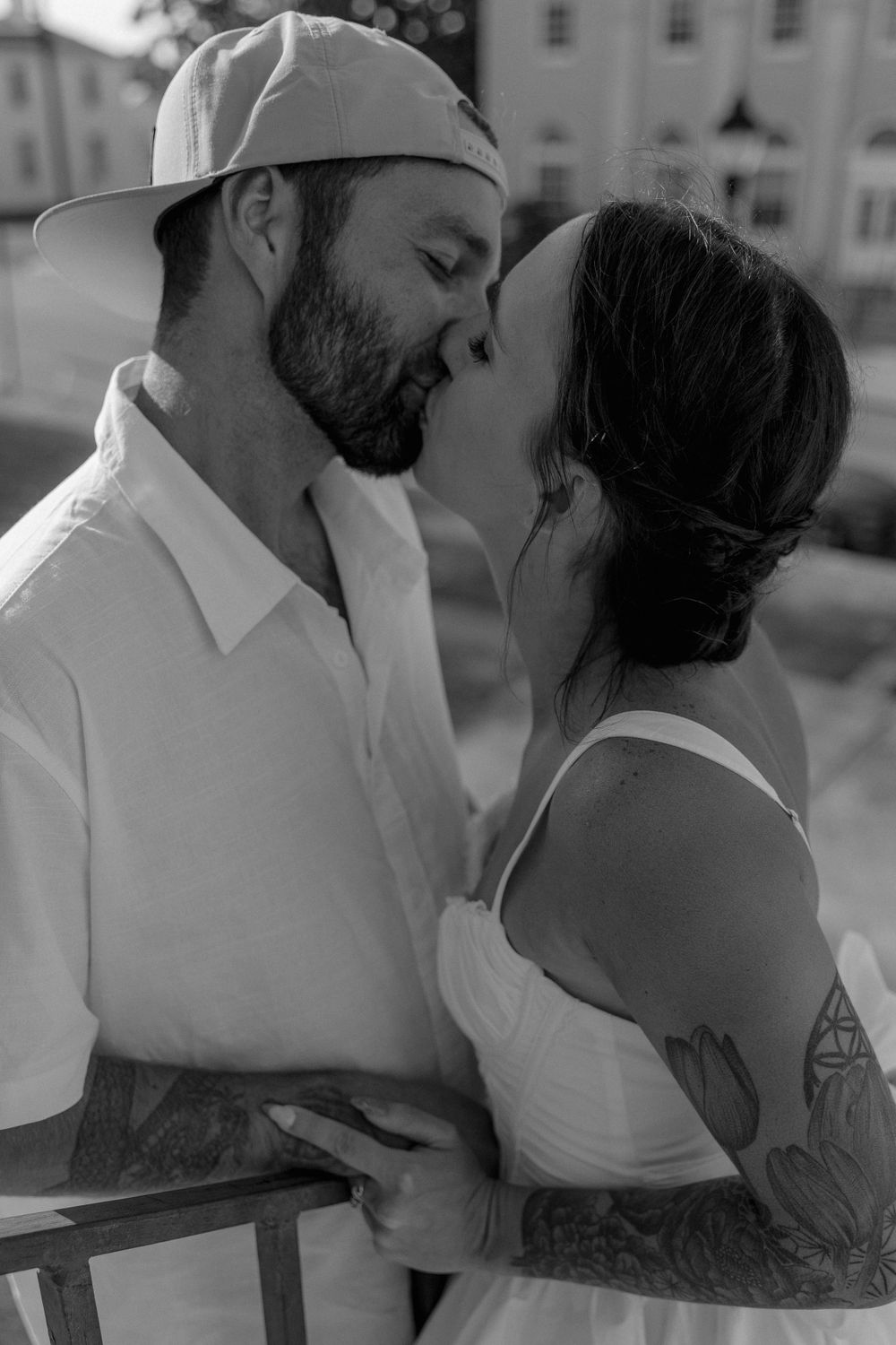 A romantic black and white portrait of two people kissing outdoors, with visible tattoos and casual summer attire.