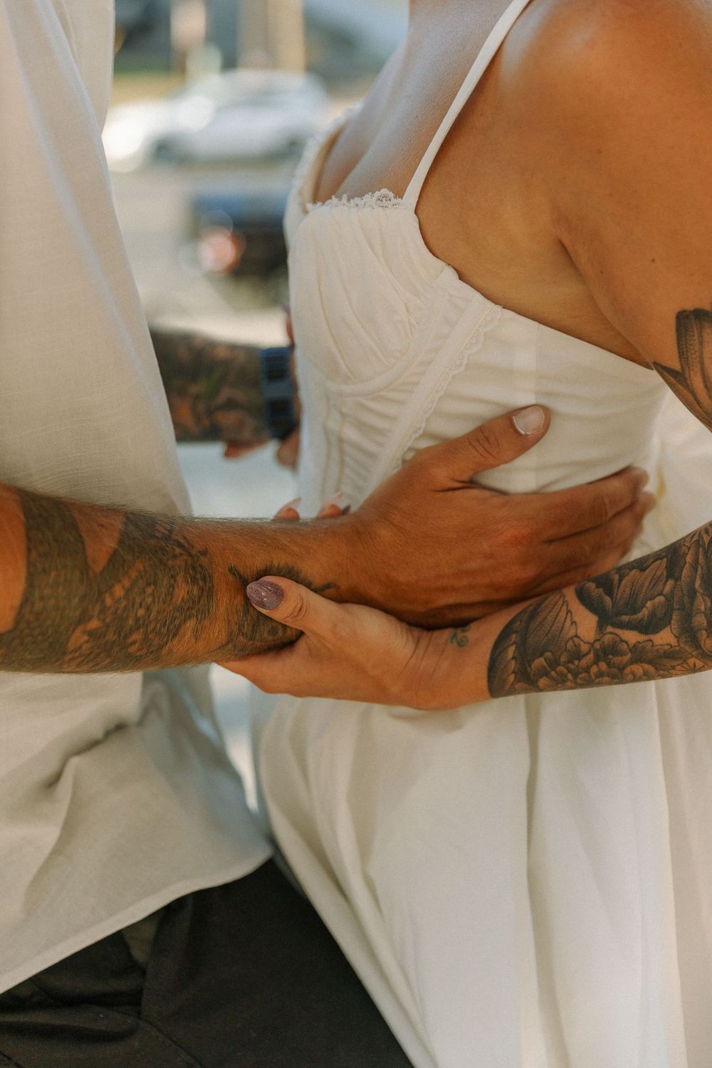Close-up of tattooed arms embracing someone wearing a white formal dress in an intimate moment.
