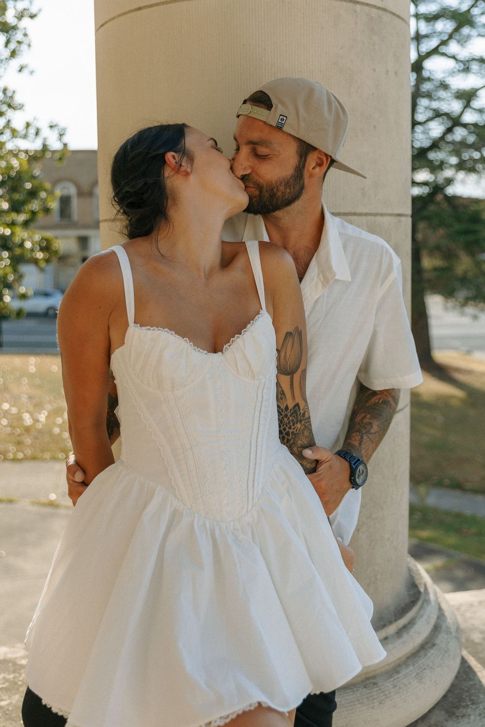 A romantic couple shares a tender kiss by a white pillar outdoors, with her in a white dress and him in casual attire.