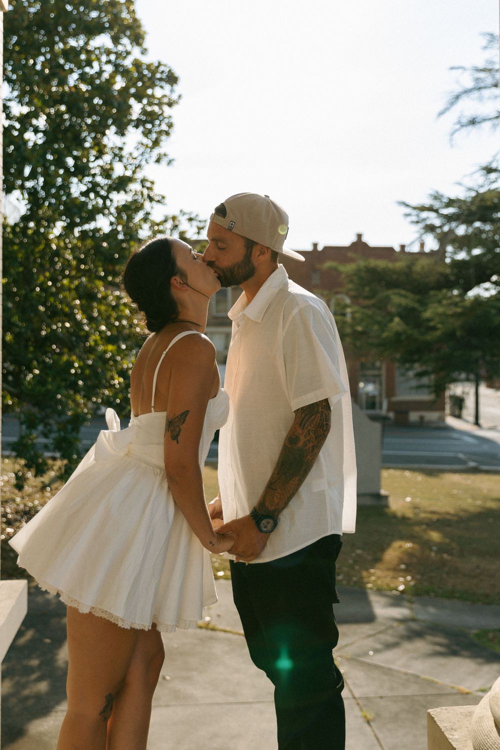 A couple in romantic attire embraces for a kiss outdoors with sunlit trees in the background.
