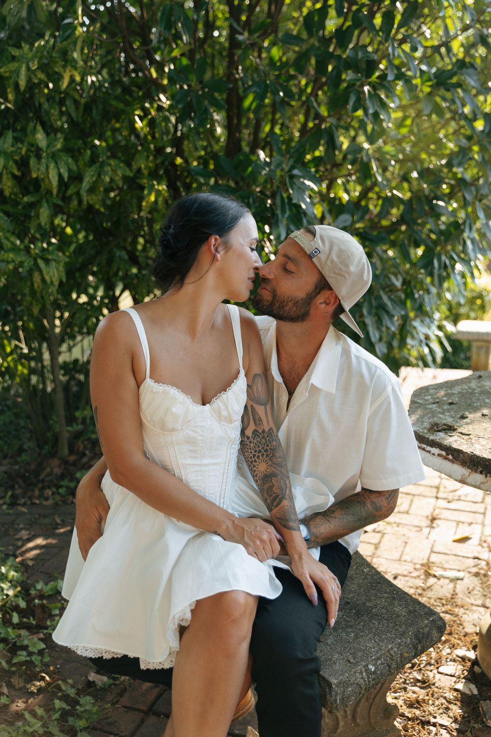 A romantic couple shares a tender moment while sitting together outdoors surrounded by lush green foliage.