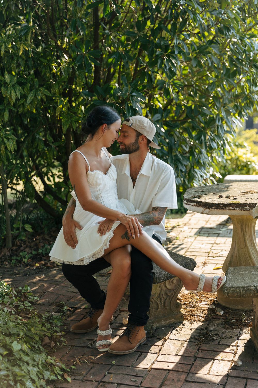 Romantic couple in white attire share tender moment while sitting on stone bench in garden setting on sunny day.