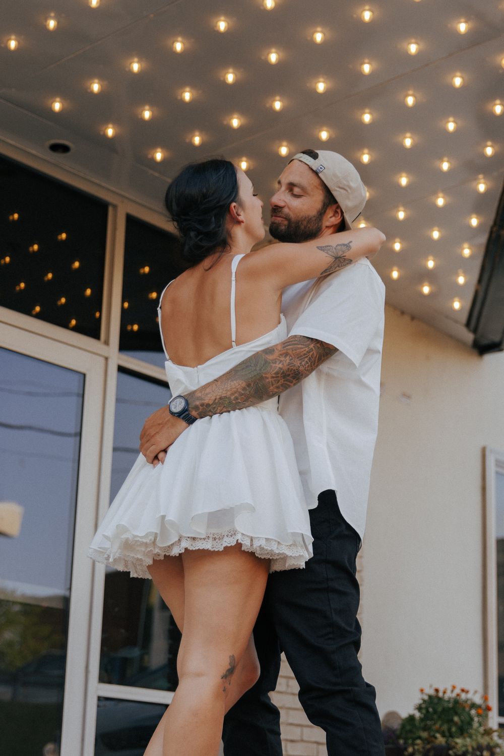 Romantic couple embraces and shares a tender moment under twinkling string lights on a porch.