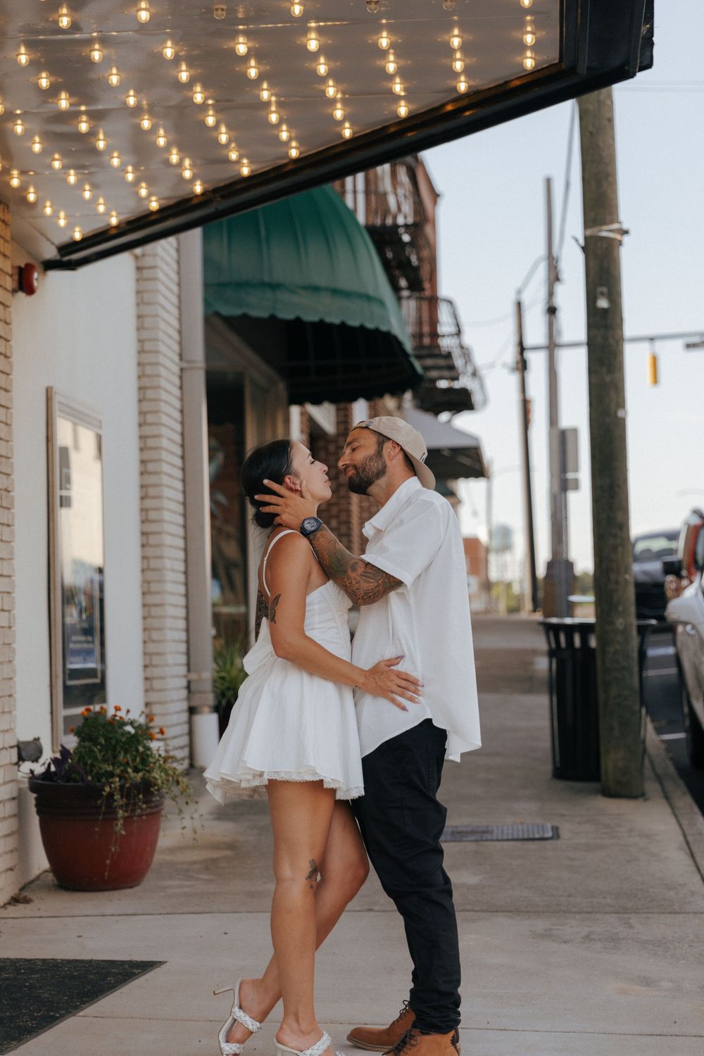 A romantic moment under string lights on a city sidewalk with white dress and casual attire.