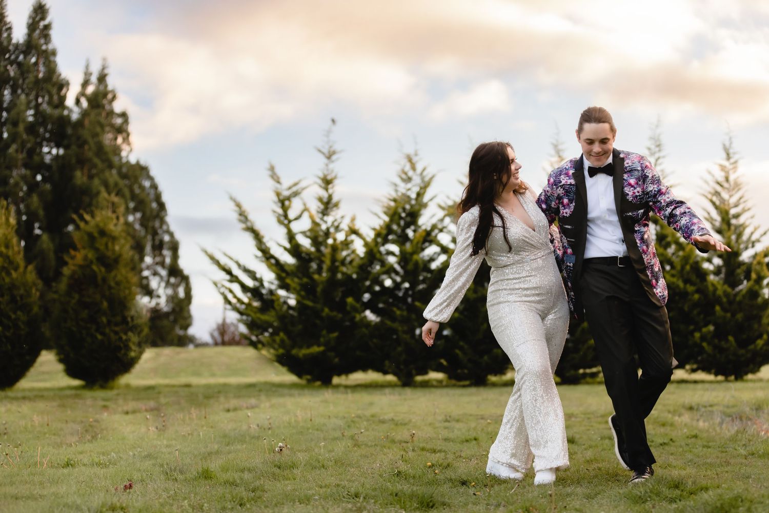 Newlywed couple walks hand-in-hand across grassy field with evergreen trees in background during golden hour.