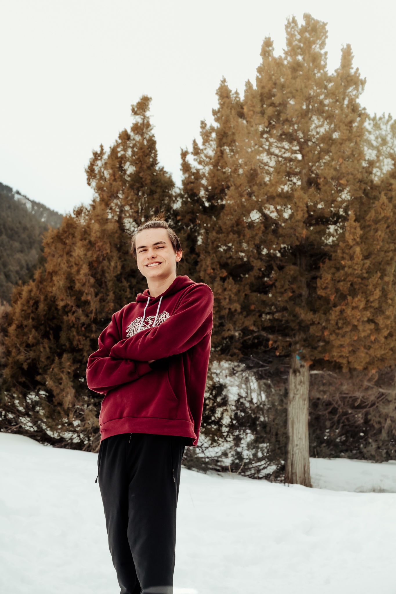 Person in burgundy hoodie posing in snowy winter setting with evergreen trees.