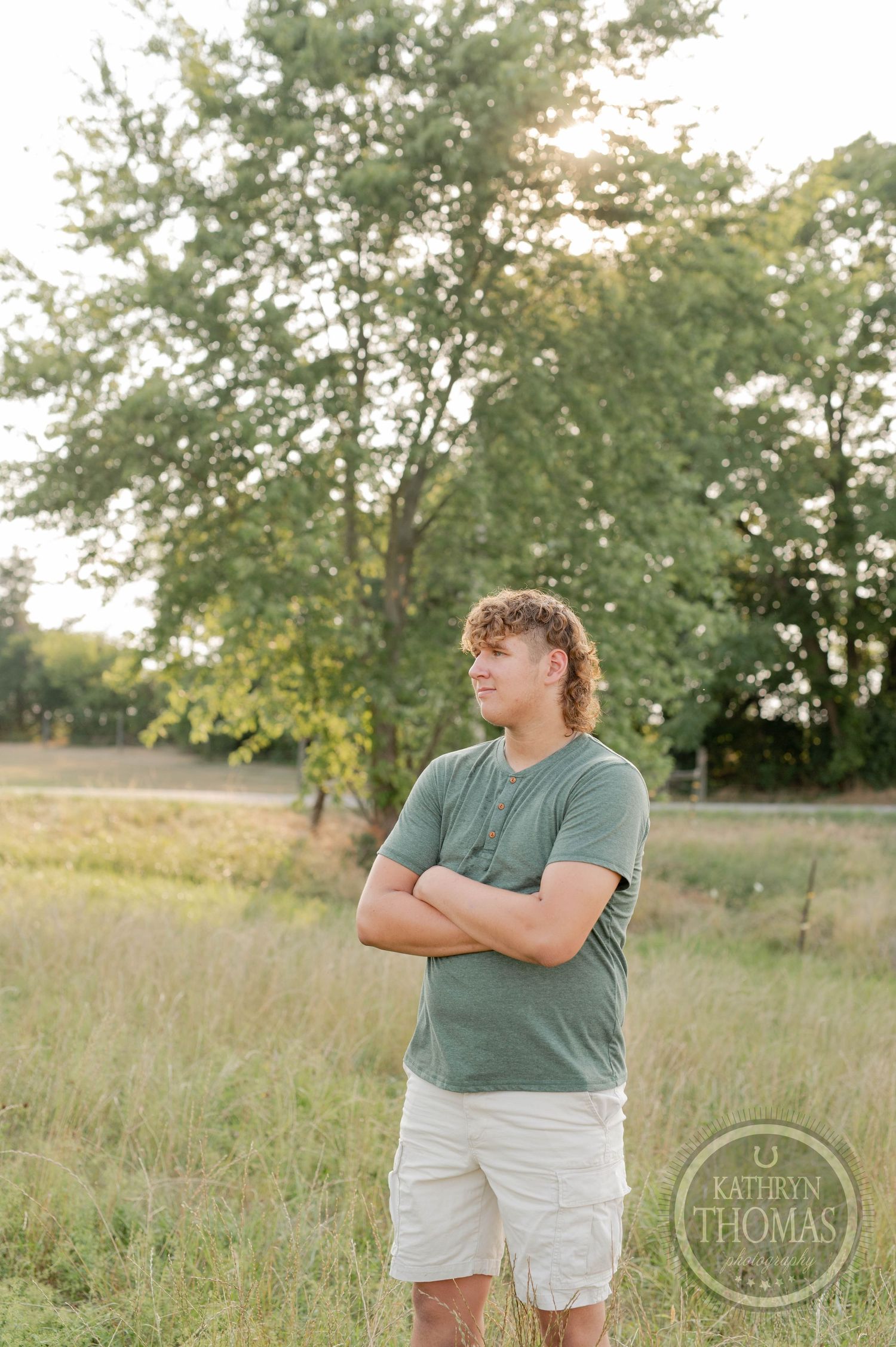 Person in sage green t-shirt stands in a grassy field with trees behind.