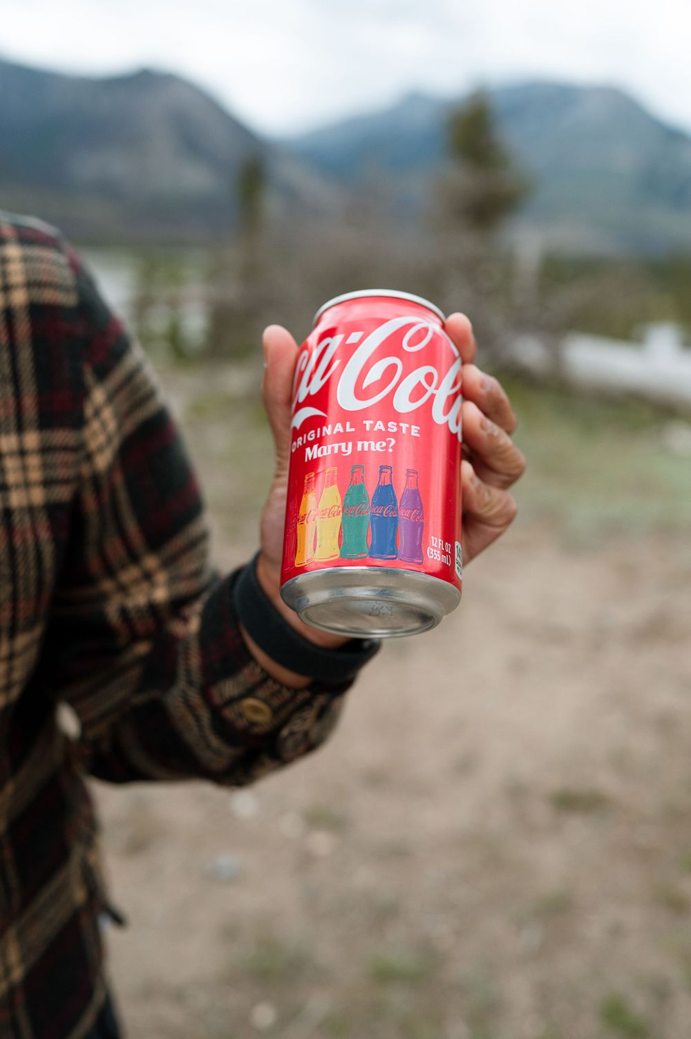 Hand holding rainbow-decorated Coca Cola can against mountain landscape.