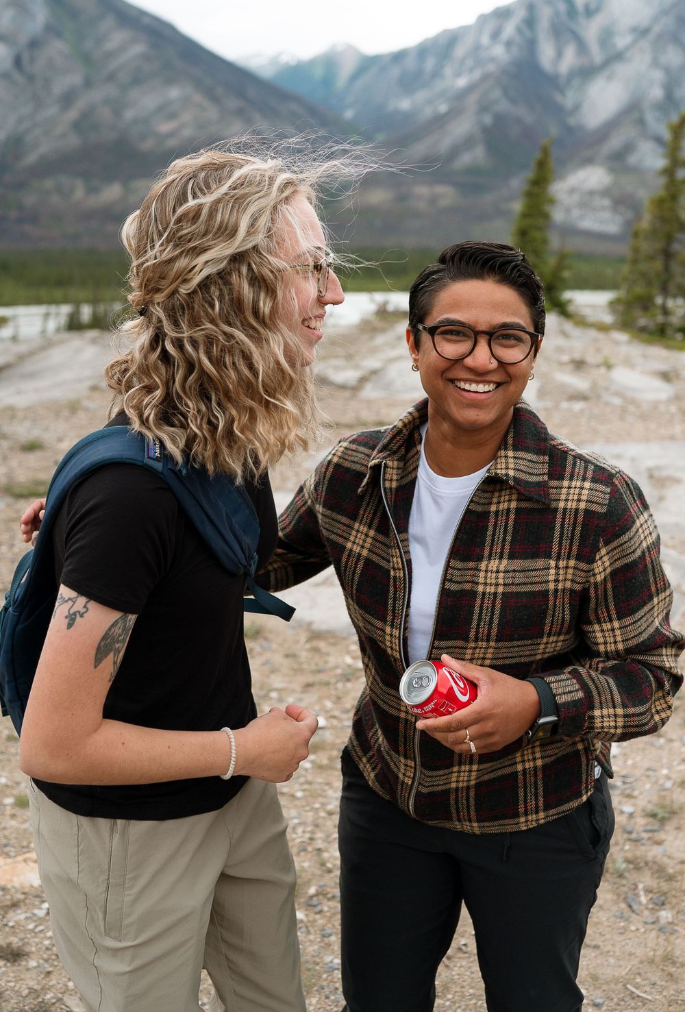 Two people smile while holding drinks against mountain scenery.