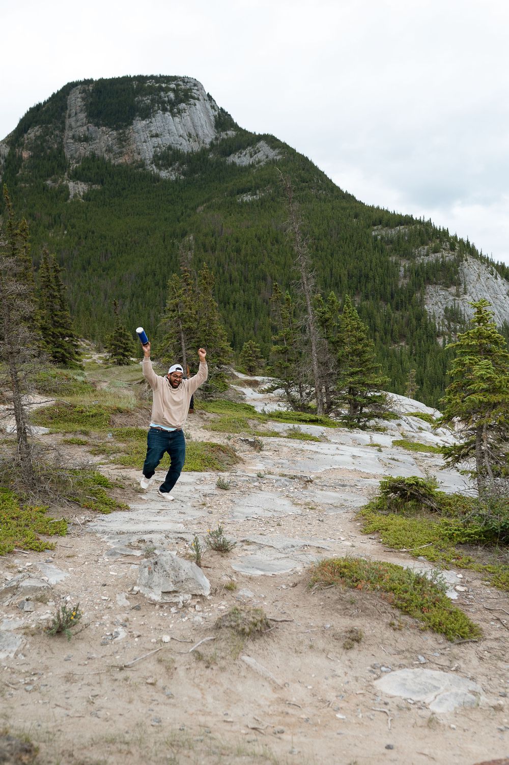 A joyful person leaps into the air on a mountain trail with evergreen trees and rocky cliffs in the background.
