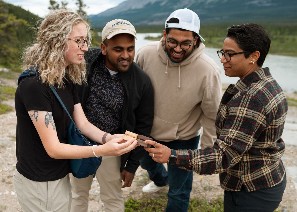 Friends gather on a mountain trail to look at something together with mountains and a lake in the background.