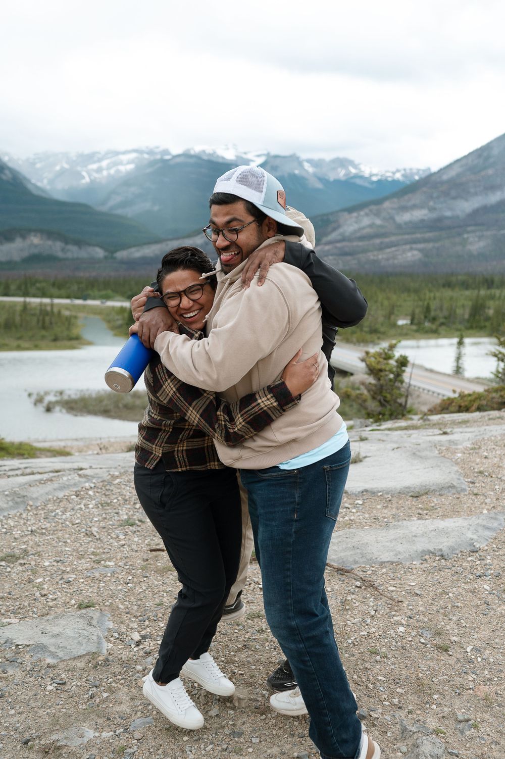 Two people embrace and laugh while standing on a rocky overlook with snow-capped mountains in the background.