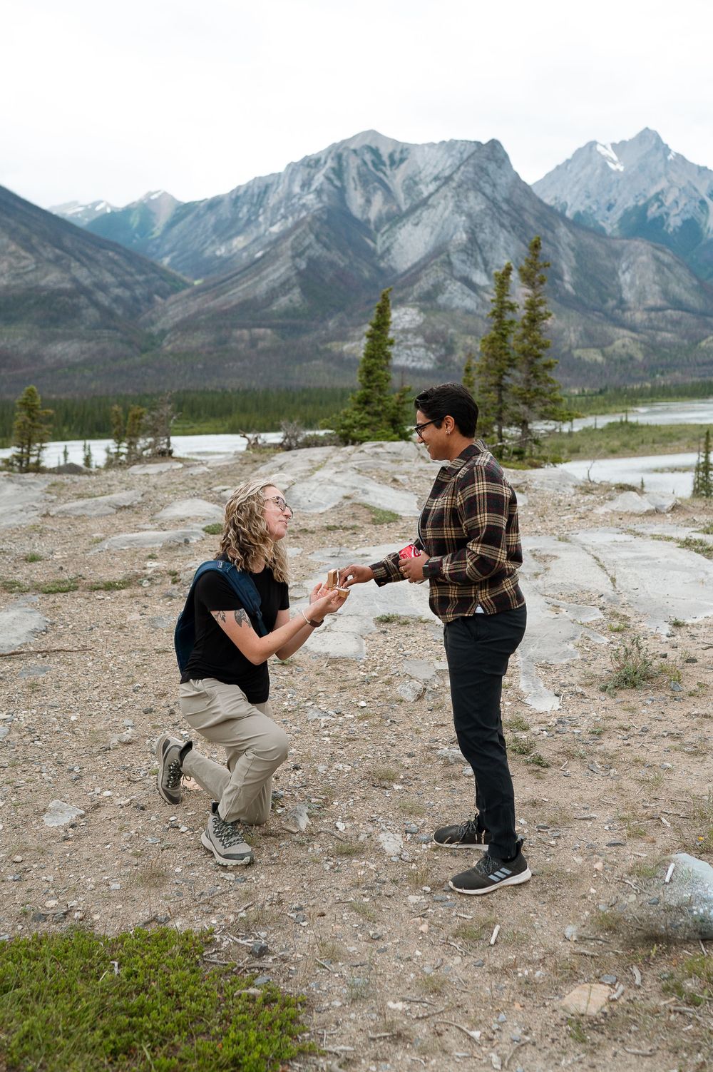 Person kneeling while proposing on rocky mountain terrain with snow-capped peaks behind.