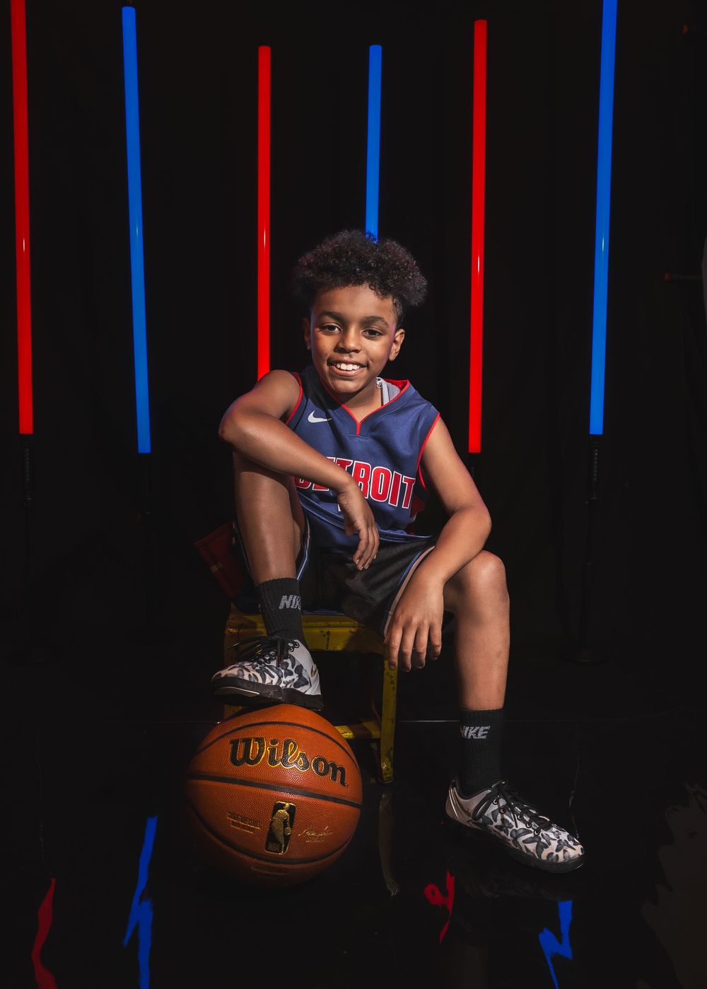 Smiling young boy in Detroit Pistons jersey poses with Wilson basketball against colorful neon light backdrop.