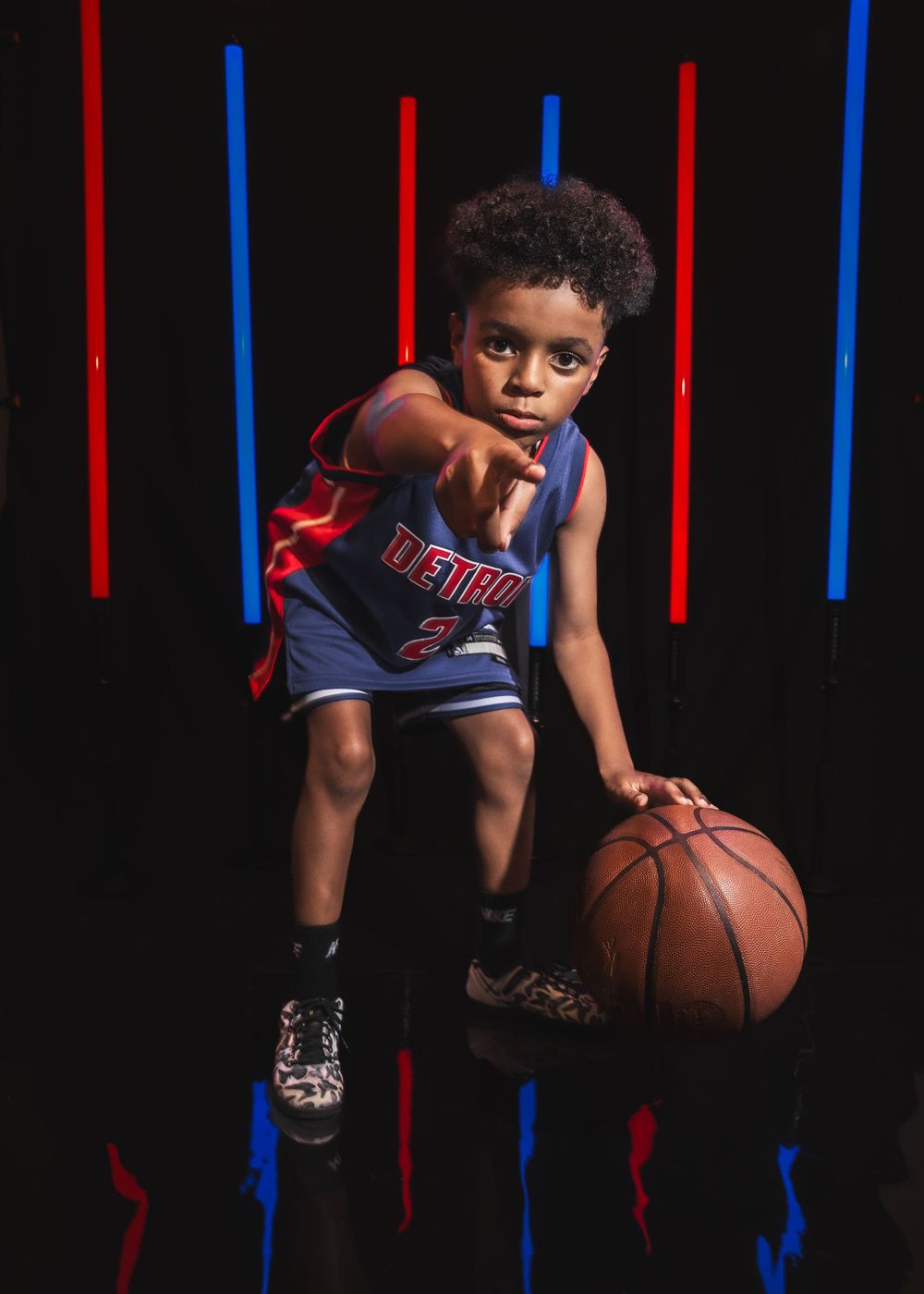 Young boy in Detroit Pistons jersey dribbling basketball, posing confidently against black background with red and blue neon lights.